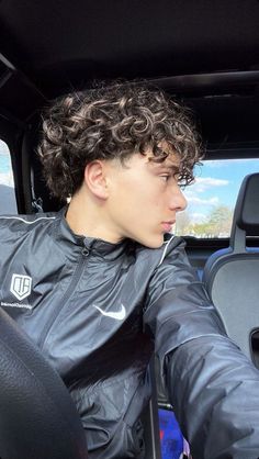 a young man sitting in the driver's seat of a car with curly hair