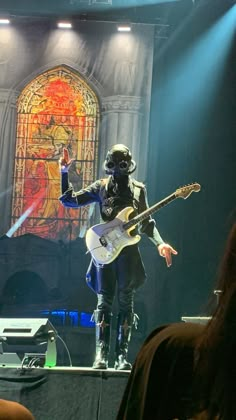 a man with a guitar on stage in front of a stained glass window and audience