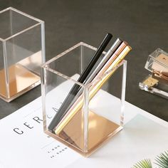 two clear boxes with pens and pencils in them sitting on a table next to a plant