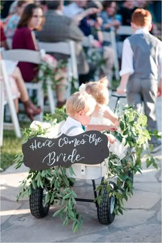 two young boys riding on a small cart with greenery and chalkboard sign that says here comes the bride