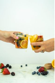 two people toasting with fruit and juice in front of sliced oranges, strawberries, and blueberries