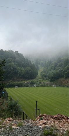 an empty soccer field surrounded by trees and hills in the distance with fog on the ground