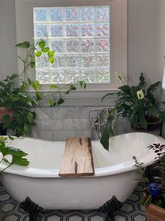 a white bath tub sitting under a window next to a plant filled bathroom sink with wooden cutting board