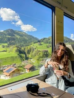 a woman sitting at a table in front of a window looking out on the countryside