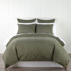 a bed with green comforter and pillows on top of wooden floor next to white wall