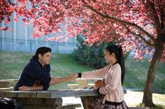 a man and woman sitting at a picnic table in front of a tree with red leaves