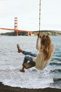 a woman sitting on a swing by the water with her feet in the air while holding onto a rope