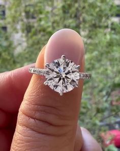 a person's hand holding a ring with a diamond in it and trees in the background