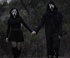 two people dressed in black holding hands and walking through the woods with scary masks on their faces