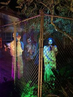 people dressed as zombies standing behind a chain link fence at night with colored lights on