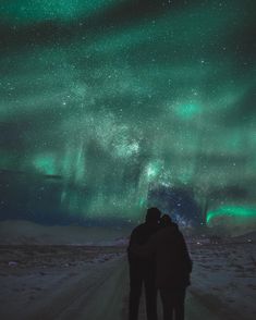 two people standing in the middle of a snow covered road under an aurora bore display