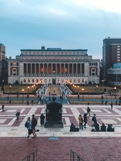 many people are walking around in front of a building with columns and lights on it