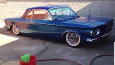 an old blue car parked in front of a brick wall with a red bucket next to it