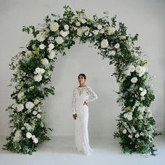 a woman standing in front of a floral arch with white roses and greenery on it