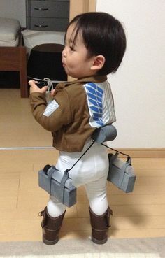 a little boy dressed in white pants and brown jacket holding a remote control while standing on the floor