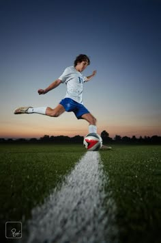 a young man kicking a soccer ball on top of a green grass covered field at sunset