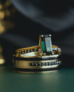 A wedding ring set from Staghead Designs sits on a dark green background. This ring set fits the alternative, edgy, gothic, nontraditional wedding aesthetic and features our "Alexa" design solitaire engagement ring in 14K yellow gold with a baguette cut sapphire center stone with knife-edge shank and a matching black diamond cuff band paired with our "Ryder" men's wedding band in 14K yellow gold with jet stone inlays and round black diamonds. Indian Winter Wedding, Alternative Couple, Black Diamond Wedding Ring, Wedding Bands Rose Gold