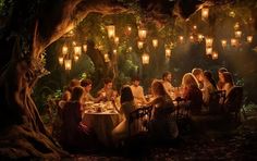 a group of people sitting around a dinner table in the woods with lanterns hanging overhead