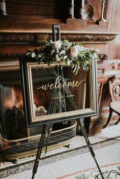 a welcome sign with flowers and greenery on it in front of a fire place