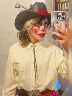 a woman in clown makeup taking a selfie with her cell phone while wearing a cowboy hat