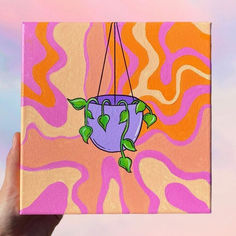 a hand holding up a painting with a plant hanging from it's center, in front of an orange and pink background