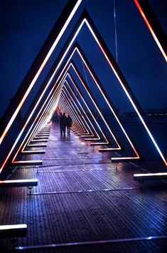 people are walking on a bridge at night with lights in the shape of triangulars