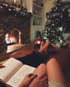 a person sitting on a couch reading a book in front of a christmas tree and fireplace