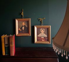 two framed portraits hang on the wall above a piano with books and a lamp next to it