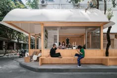 three people sitting on a wooden bench in front of a building with white roof and windows