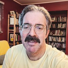 a man with glasses and a mustache is sitting in front of a bookshelf