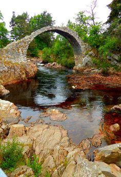 an old stone bridge over a river surrounded by rocks and greenery in the foreground