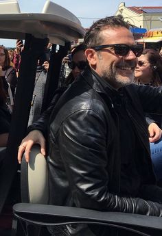 a man in black leather jacket and sunglasses sitting on a bus with people behind him