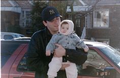 a man holding a small child in his arms next to a red car and trees