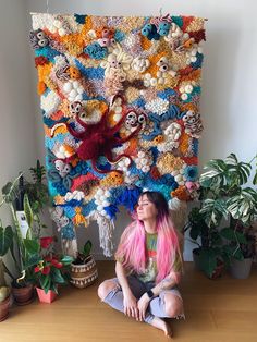 a woman with pink hair sitting on the floor in front of a colorful wall hanging