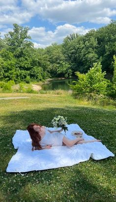 a woman laying on top of a white blanket in the grass next to a lake