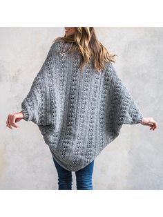 a woman standing in front of a concrete wall wearing a gray knitted ponchy