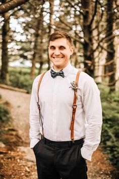 a man wearing a bow tie and suspenders standing in the woods with his hands in his pockets