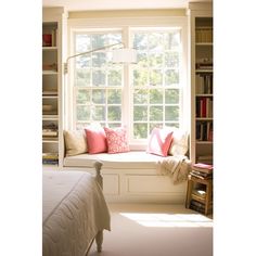 a bed sitting under a window next to a white book shelf filled with books and pillows