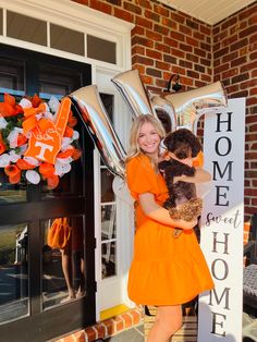 a woman in an orange dress is holding a teddy bear and some balloons on the front porch