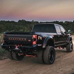 a large black truck driving down a dirt road next to a lush green forest at sunset