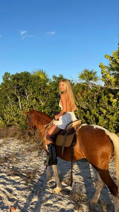 a woman riding on the back of a brown horse down a sandy beach next to trees