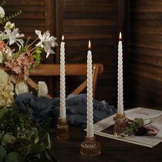 three white candles sitting on top of a wooden table next to flowers and a book