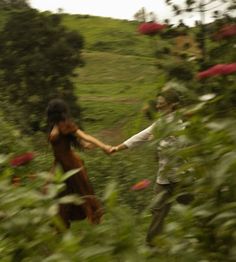 two people holding hands in the middle of a lush green field with red flowers on it