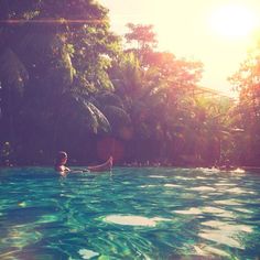 a person in a pool with trees and sun shining on the water behind them,