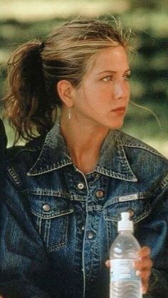 a woman in denim jacket holding a water bottle and looking off into the distance with trees behind her