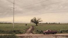 an old red truck parked on the side of a dirt road next to a field