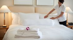 a woman unpacking white sheets on a bed with folded towels and flower in the foreground