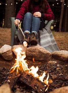 a woman sitting in a chair next to a campfire with her feet on the ground