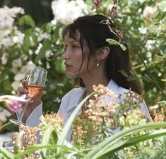 a woman holding a glass of wine in her hand and looking at the flowers behind her