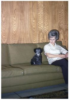 an older woman sitting on a couch next to a small black dog in front of a wood paneled wall